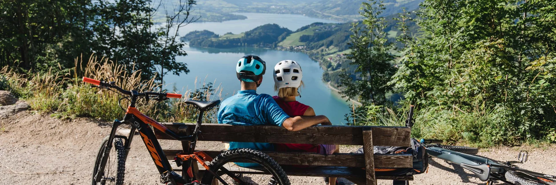 Twee fietsers zitten op een bankje met uitzicht op een meer en bergen.