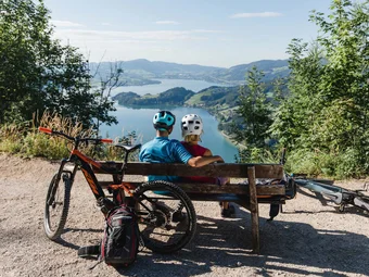 Two cyclists sitting on a bench overlooking a lake and mountains.