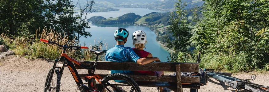 Die besten Touren & Trails der Alpen © Katrin Kerschbaumer Zwei Radfahrer sitzen auf einer Bank und blicken auf einen See und Berge.