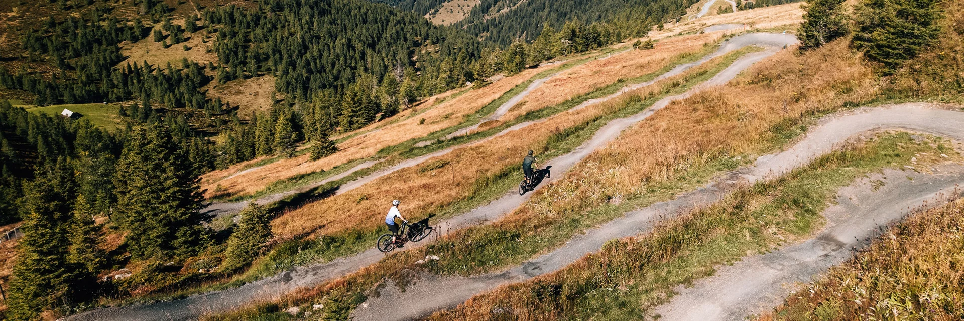 Zwei Radfahrer auf einem kurvigen Bergweg umgeben von Wäldern und Bergen