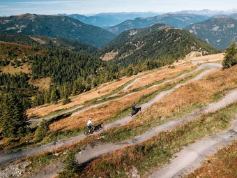 Two cyclists on a winding mountain trail surrounded by forests and mountains