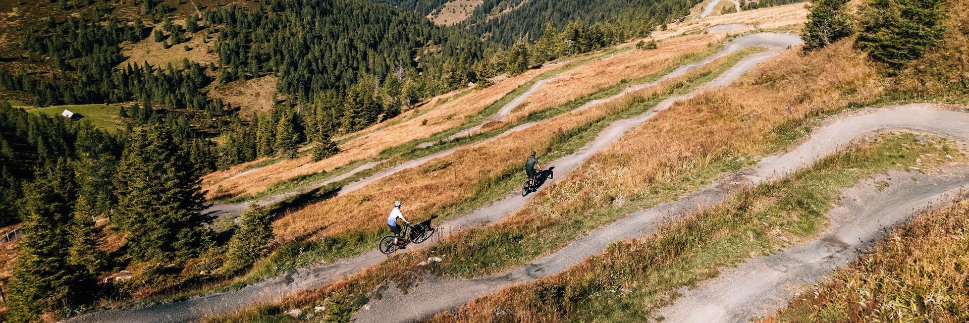 Zwei Radfahrer auf einem kurvigen Bergweg umgeben von Wäldern und Bergen