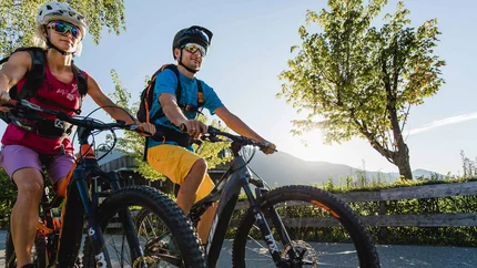Zwei Radfahrer mit Schutzhelm fahren auf Landstraße bei sonnigem Wetter