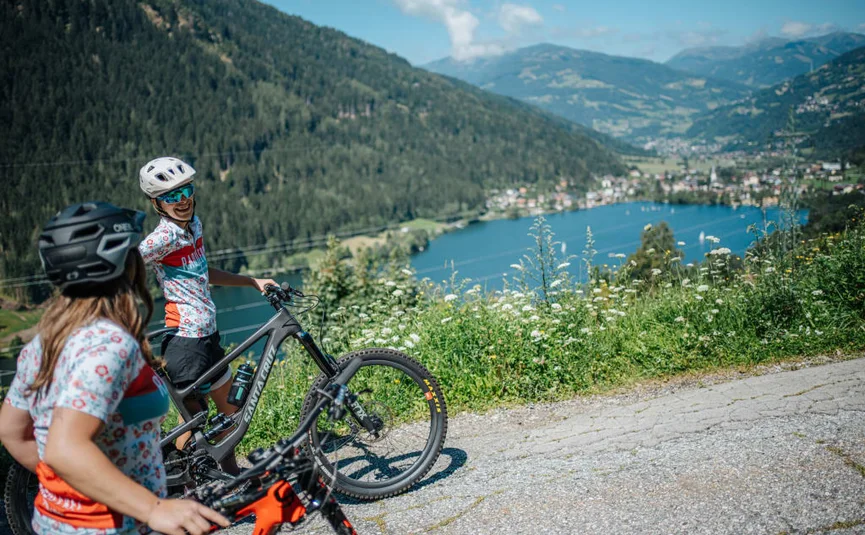 Two cyclists with helmets on mountain path overlooking lake and mountains