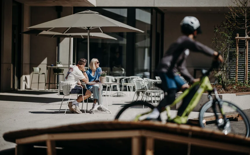 Koppel zit buiten aan tafel onder parasol, fietser rijdt voorbij