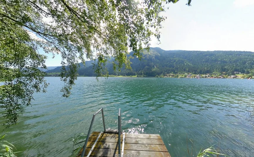 View of a lake with a wooden dock, forested hills, and sailboats in the distance