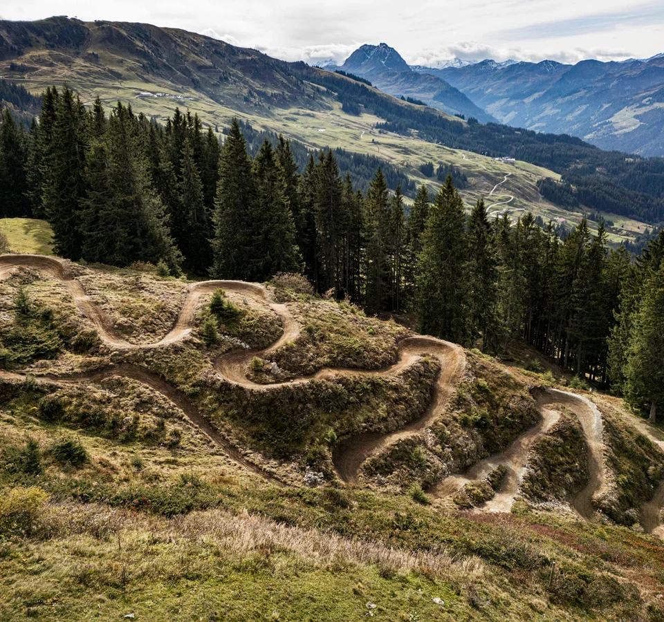 Neuer Flow im Brixental: Der Kirchberg Trail eröffnet © Stromberg Schmaler kurviger Mountainbike-Trail auf Berghang mit Tannen und Bergen im Hintergrund