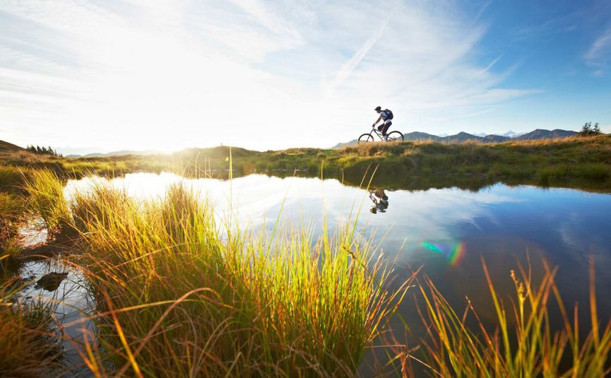 BIKE BREAK (4 nights) Cyclist riding on trail beside lake at sunset with mountains in background