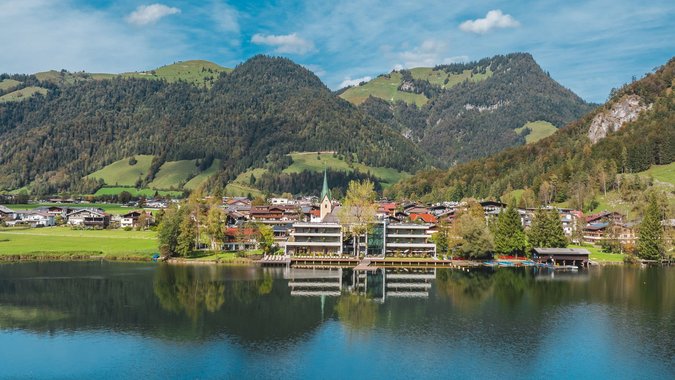 Bergdorf am See mit bewaldeten Hügeln und blauen Himmel im Hintergrund