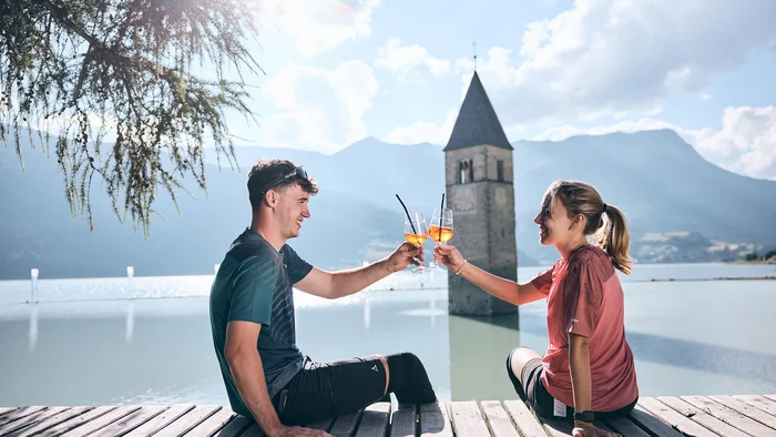 Junges Paar stößt mit Aperol am gefluteten Kirchturm in Reschen an.