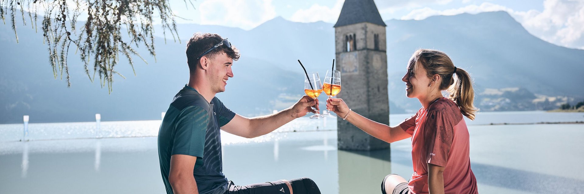 Alpen-Comfort-Hotel Central **** © Tobias Köhler Young couple toasting with Aperol by flooded church tower in Reschen.