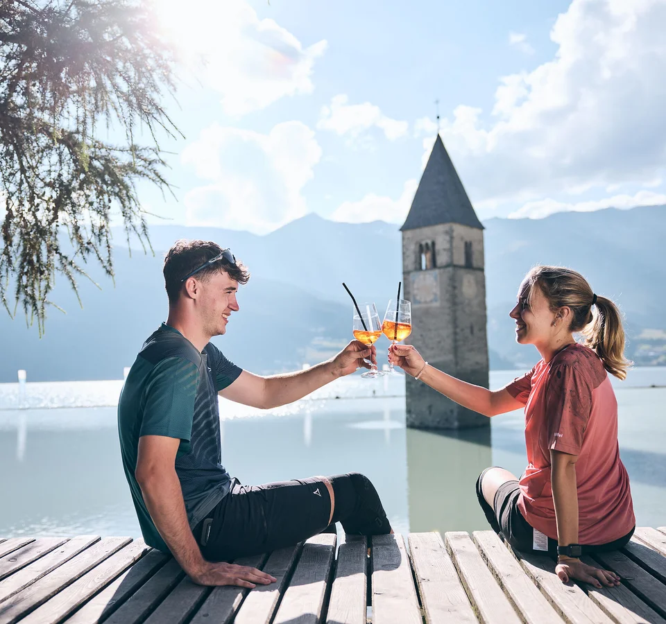 Junges Paar stößt mit Aperol am gefluteten Kirchturm in Reschen an.