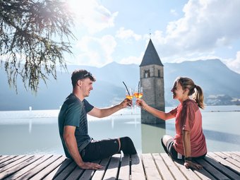 Young couple toasting with Aperol by flooded church tower in Reschen.
