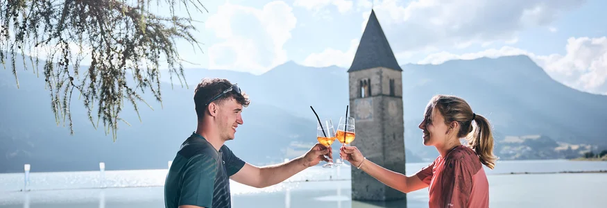 Junges Paar stößt mit Aperol am gefluteten Kirchturm in Reschen an.