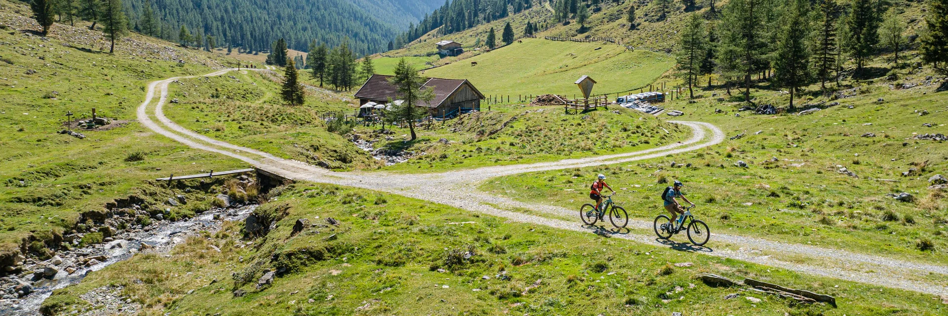 Two cyclists riding on a mountain trail through green alpine scenery