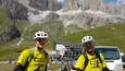 Zwei Radfahrer in gelben Trikots vor Bergkulisse und Autos