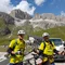 Zwei Radfahrer in gelben Trikots vor Bergkulisse und Autos