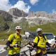 Zwei Radfahrer in gelben Trikots vor Bergkulisse und Autos
