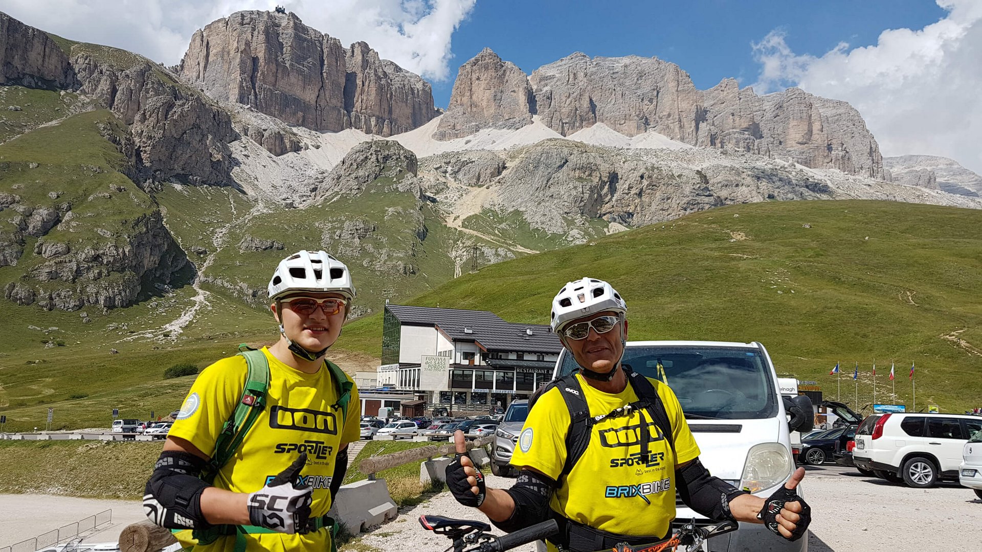 Zwei Radfahrer in gelben Trikots vor Bergkulisse und Autos