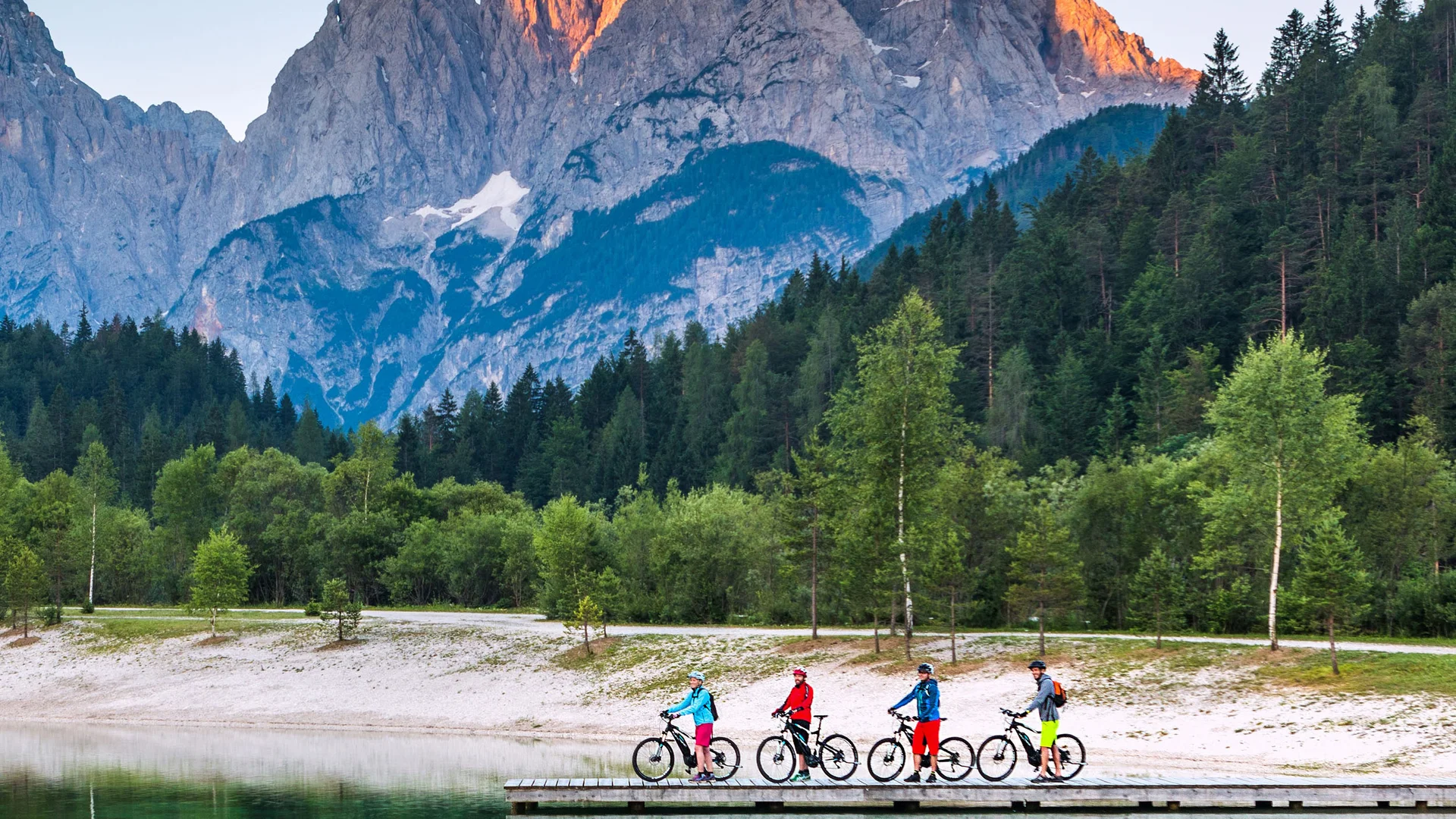Vier Radfahrer stehen auf einem Steg am See vor Bergen im Abendlicht