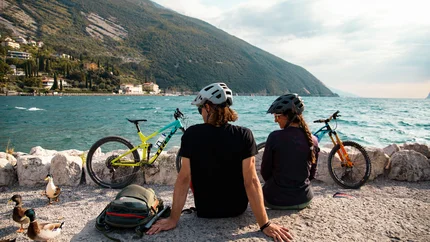 Zwei Fahrradfahrer mit Helmen sitzen am See mit Blick auf Berge und Wasser