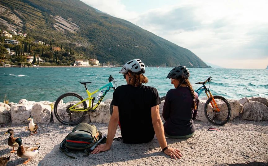 Two cyclists wearing helmets sitting by a lake with mountains and water view