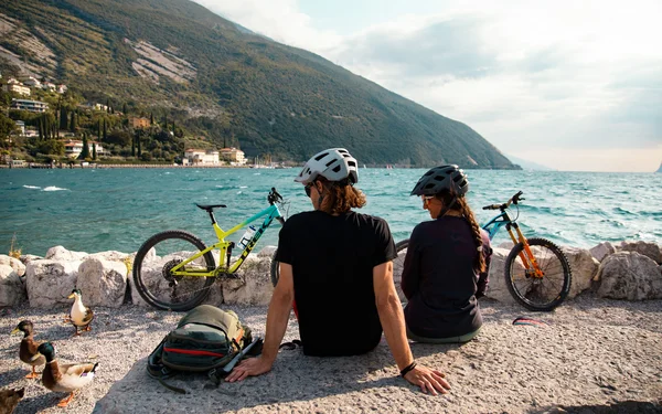 Zwei Fahrradfahrer mit Helmen sitzen am See mit Blick auf Berge und Wasser