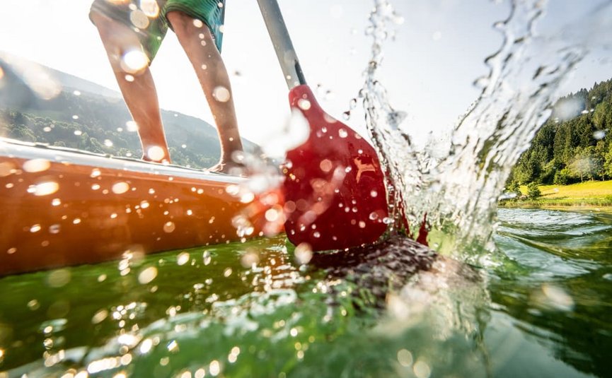 Person rudert mit rotem Paddel auf grünem See bei sonnigem Wetter