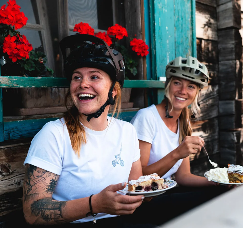 Ein perfekter Enduro-Tag – vom ersten Höhenmeter bis zum letzten Trail © saalbach.com, Moritz Ablinger Zwei lachende Frauen mit Fahrradhelmen essen Kuchen vor einem Holzhaus mit roten Blumen