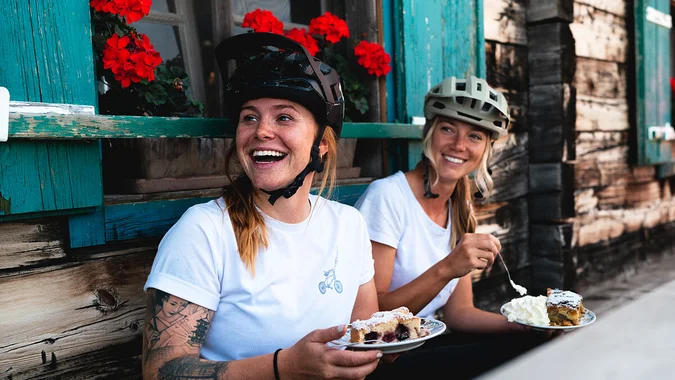 Mountain Bike Holidays © saalbach.com, Moritz Ablinger Zwei lachende Frauen mit Fahrradhelmen essen Kuchen vor einem Holzhaus mit roten Blumen