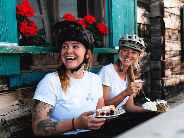 Happy Birthday EPIC Bike Park! © saalbach.com, Moritz Ablinger Zwei lachende Frauen mit Fahrradhelmen essen Kuchen vor einem Holzhaus mit roten Blumen