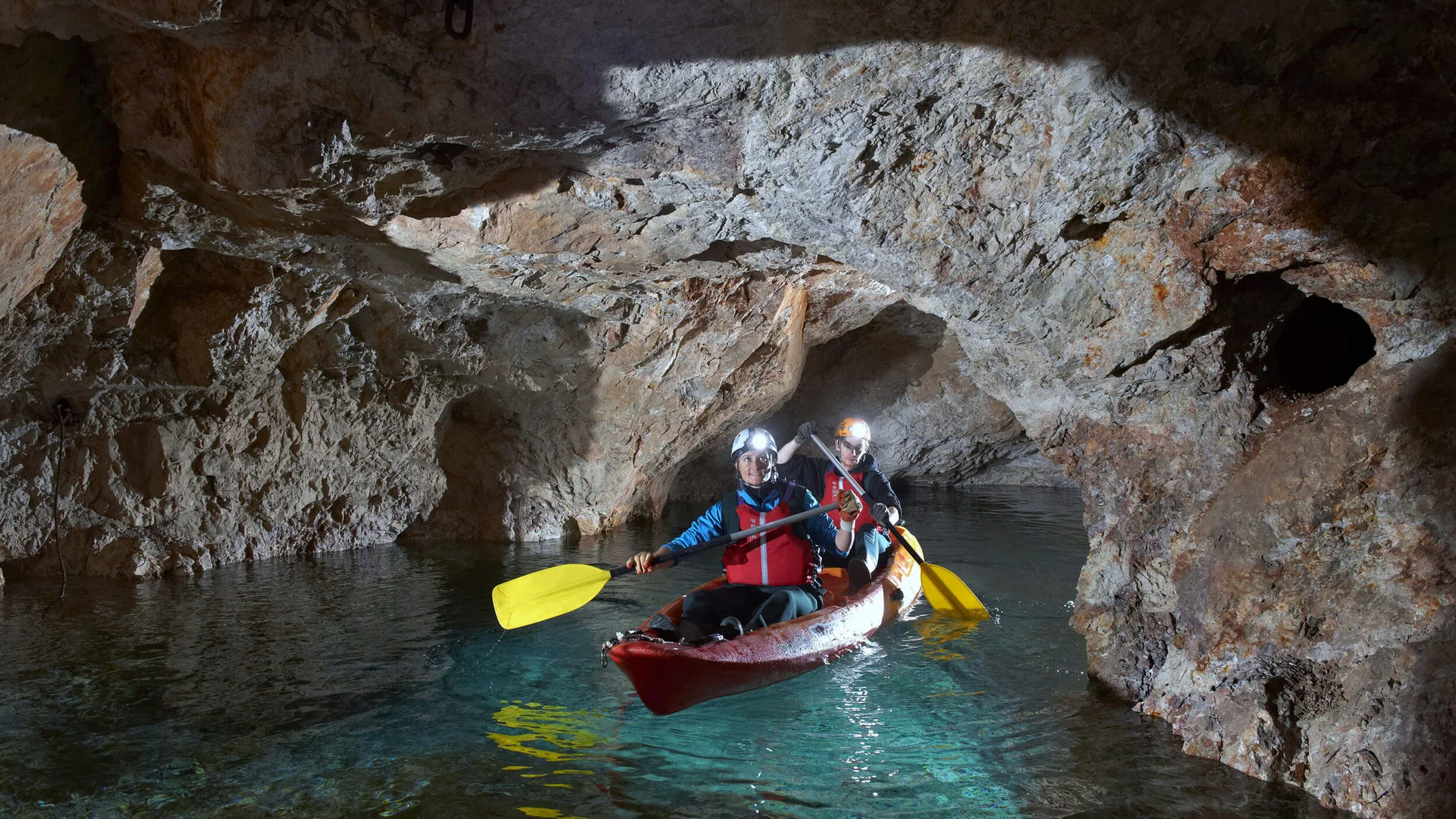 Zwei Personen paddeln in einem Kajak durch eine beleuchtete Höhle auf klarem Wasser