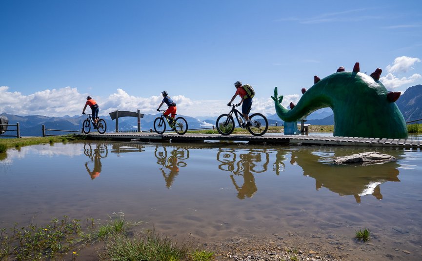 Drei Mountainbiker auf Holzbrücke neben grüner Dinosaurierstatue am Teich
