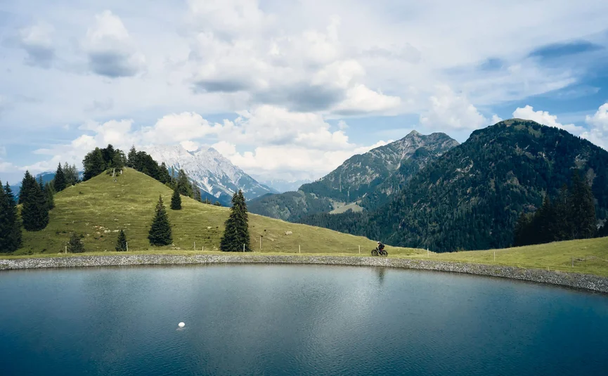 Fahrradfahrer auf einem Weg neben einem Bergsee mit Bergen im Hintergrund