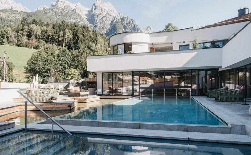 Moderner Pool mit Liegestühlen vor Bergkulisse und weißem Gebäude