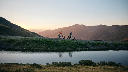 Zwei Mountainbiker fahren bei Sonnenuntergang auf einem Bergweg neben einem See