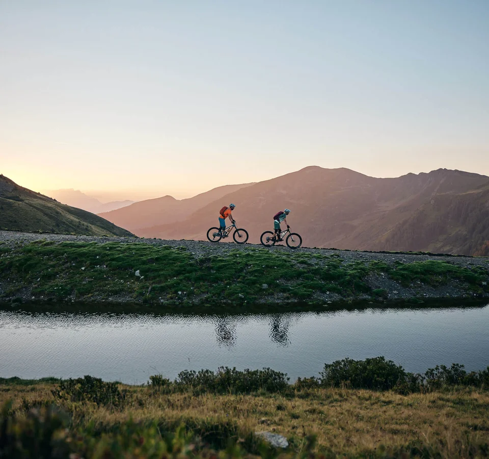 Unterwegs in Österreichs größter Bikeregion © Tobias Köhler Zwei Mountainbiker fahren bei Sonnenuntergang auf einem Bergweg neben einem See