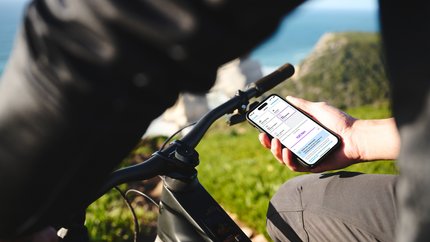 Person hält Smartphone mit Fahrrad-App neben Fahrradlenker im Freien