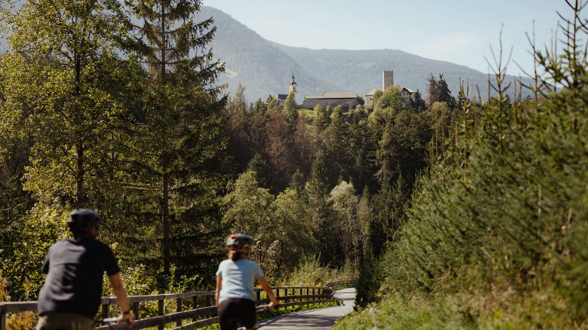 Zwei Radfahrer auf einem Waldweg mit einem Schloss und Bergen im Hintergrund
