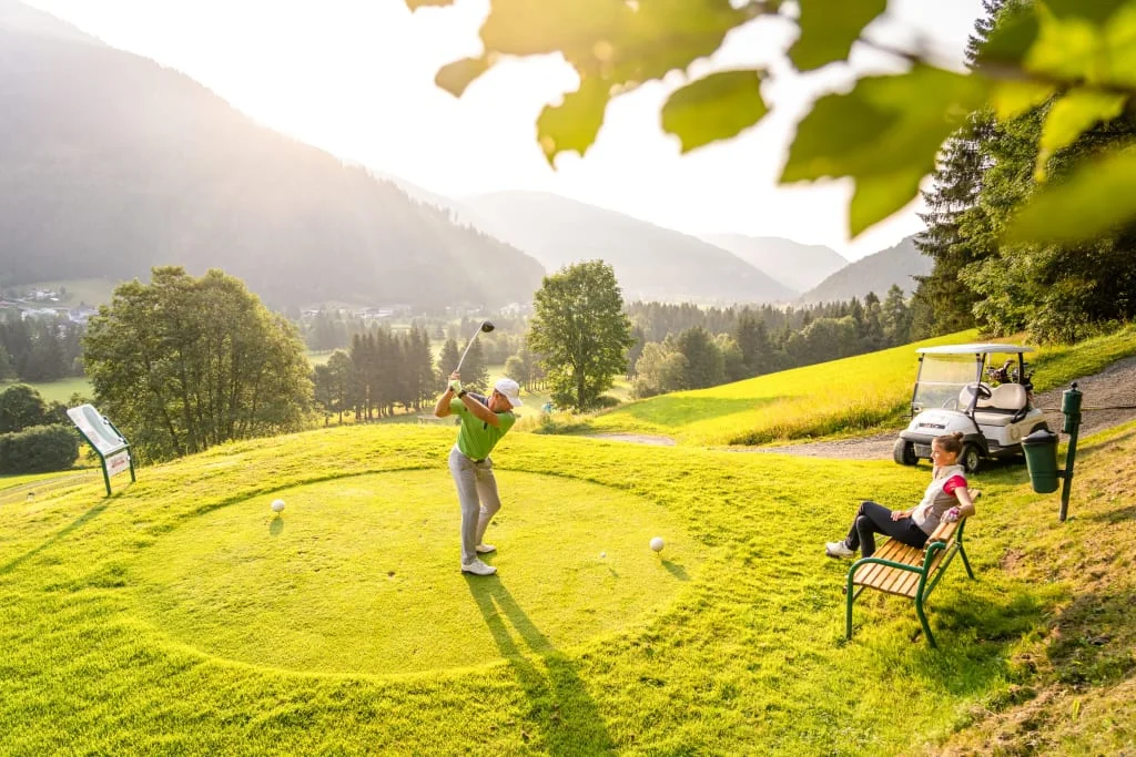 Golfer hitting ball on sunny mountain golf course with caddy sitting