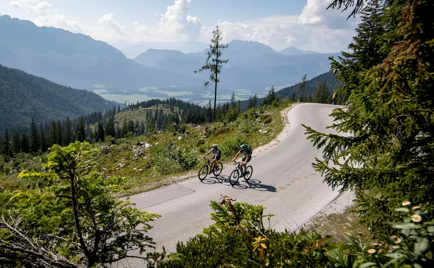 Two cyclists riding on mountain road with forest and mountains in background