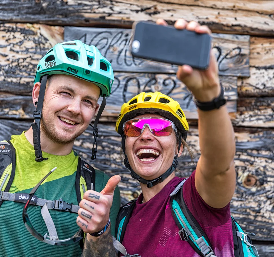 Ein perfektes Bike-Wochenende in Ischgl © WOM Medien - Andreas Meyer Zwei Fahrradfahrer machen Selfie vor Holzwand, lächelnd und Daumen hoch