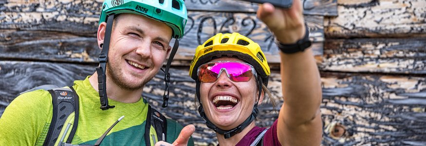 Zwei Fahrradfahrer machen Selfie vor Holzwand, lächelnd und Daumen hoch
