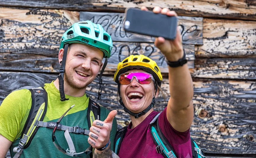 Zwei Fahrradfahrer machen Selfie vor Holzwand, lächelnd und Daumen hoch