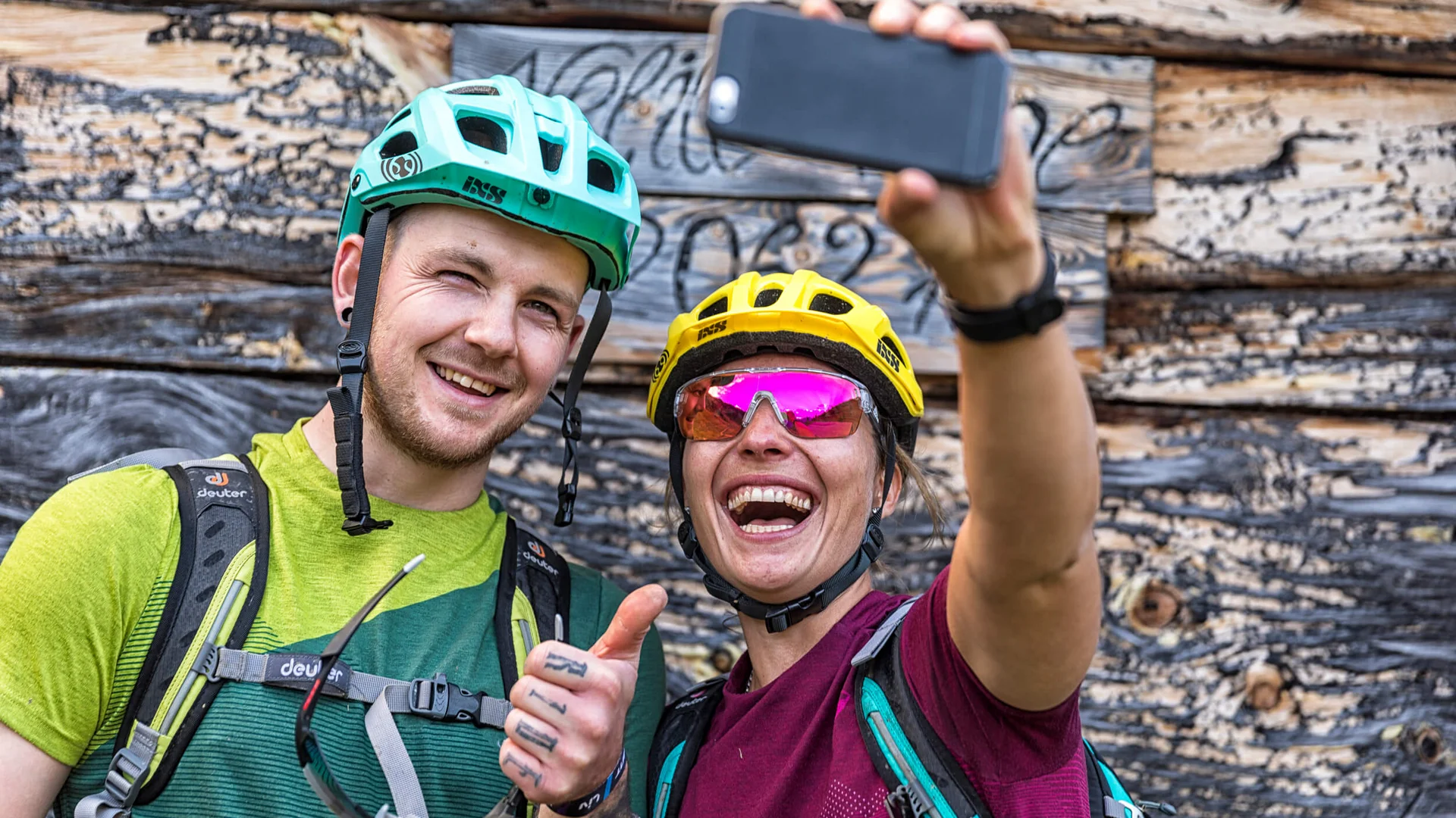 Wir sind E-Bike. © WOM Medien - Andreas Meyer Zwei Fahrradfahrer machen Selfie vor Holzwand, lächelnd und Daumen hoch