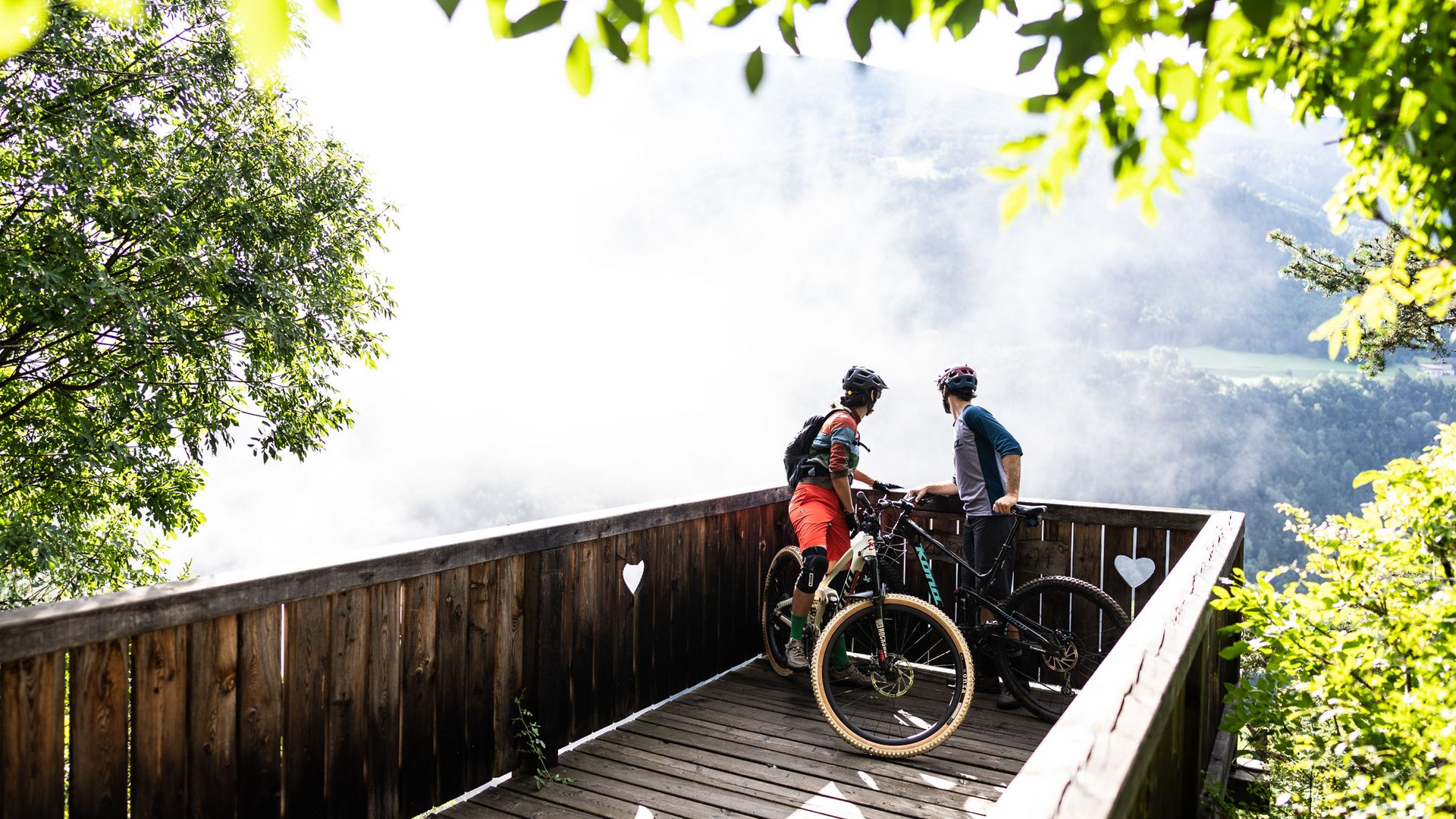 Zwei Mountainbiker auf einer Aussichtsplattform schauen ins neblige Tal