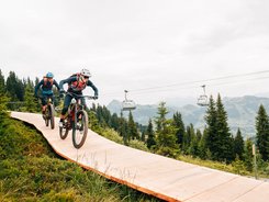 Zwei Mountainbiker fahren bergab auf einem Holzweg umgeben von Tannenbäumen