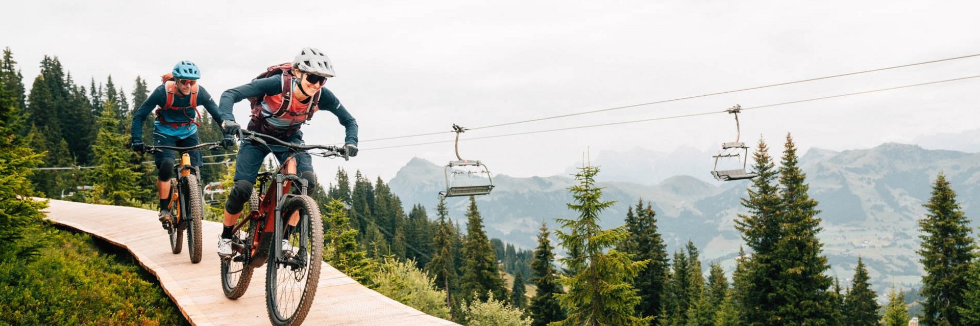 Zwei Mountainbiker fahren bergab auf einem Holzweg umgeben von Tannenbäumen
