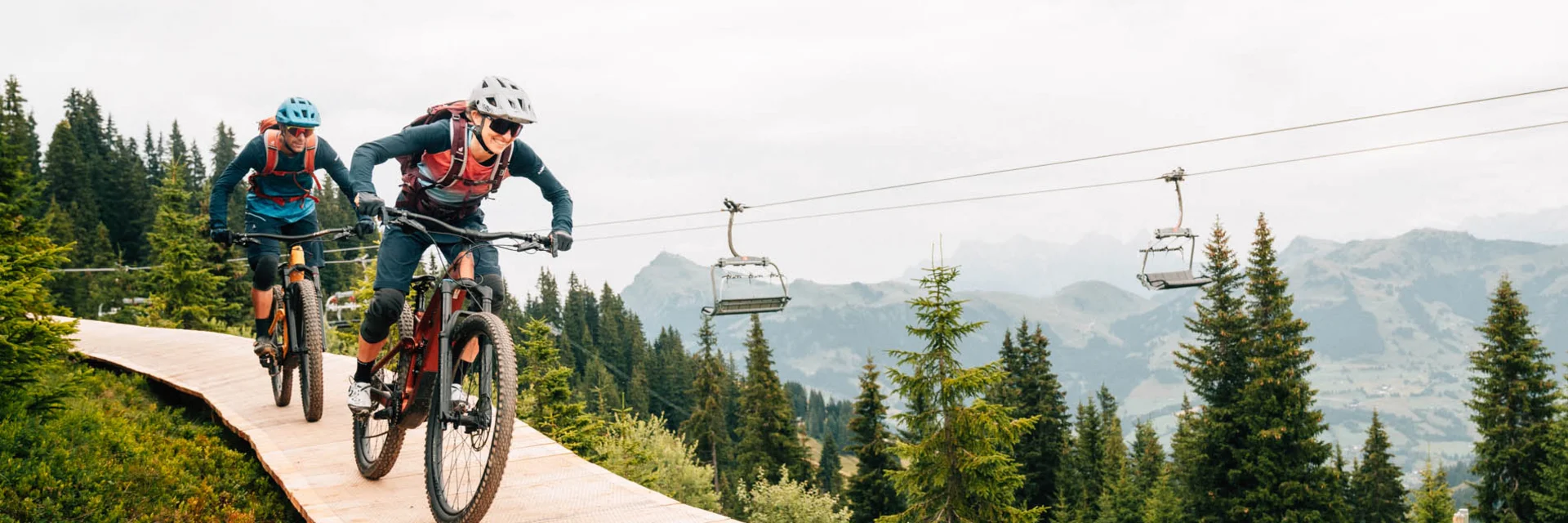 Zwei Mountainbiker fahren bergab auf einem Holzweg umgeben von Tannenbäumen