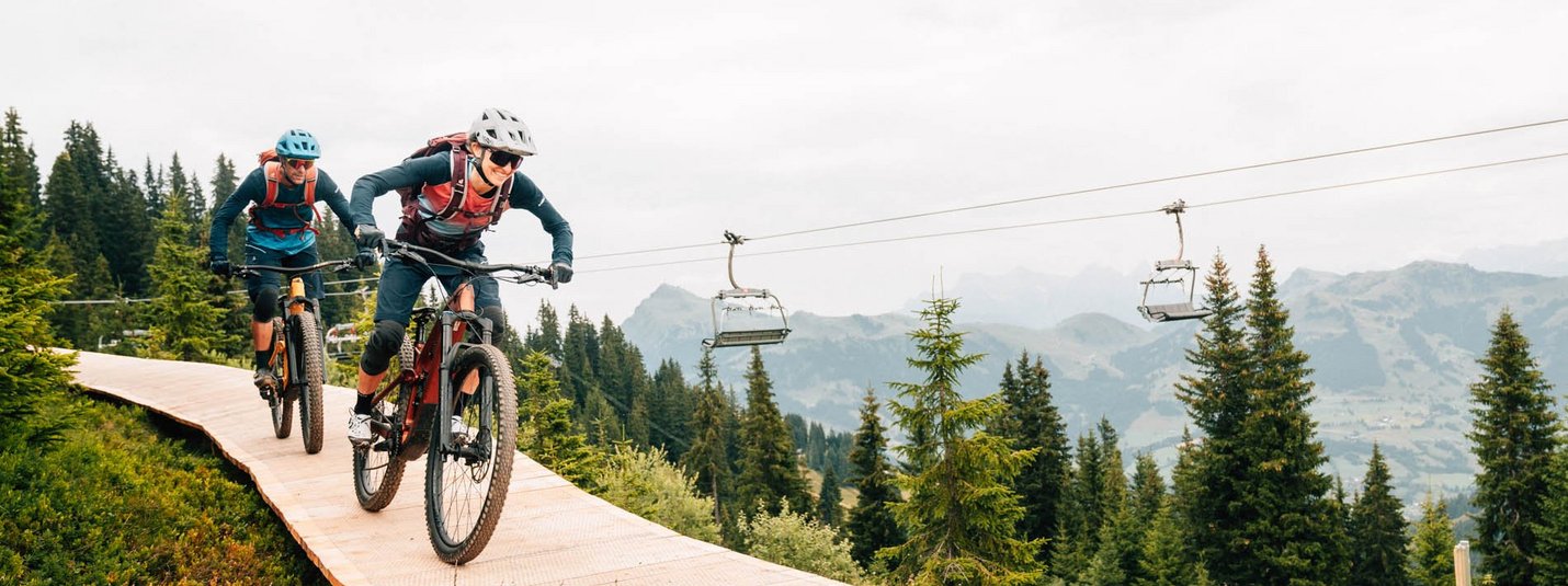 Zwei Mountainbiker fahren bergab auf einem Holzweg umgeben von Tannenbäumen